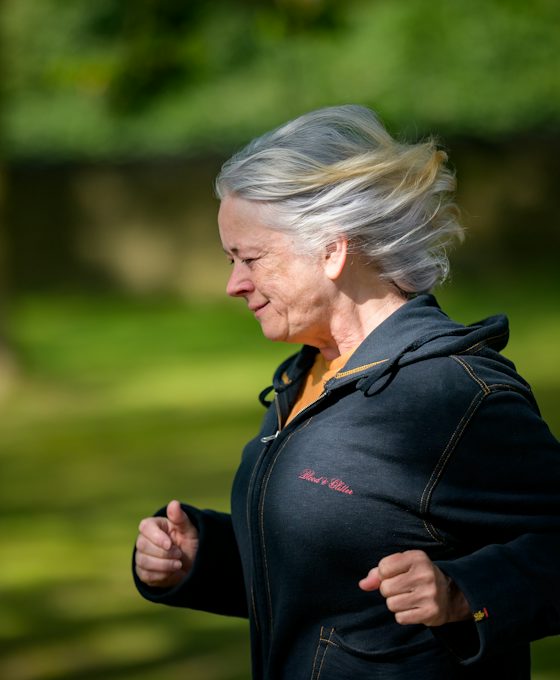 A smiling woman in her sixties with silver hair running outdoors in a sunlit park